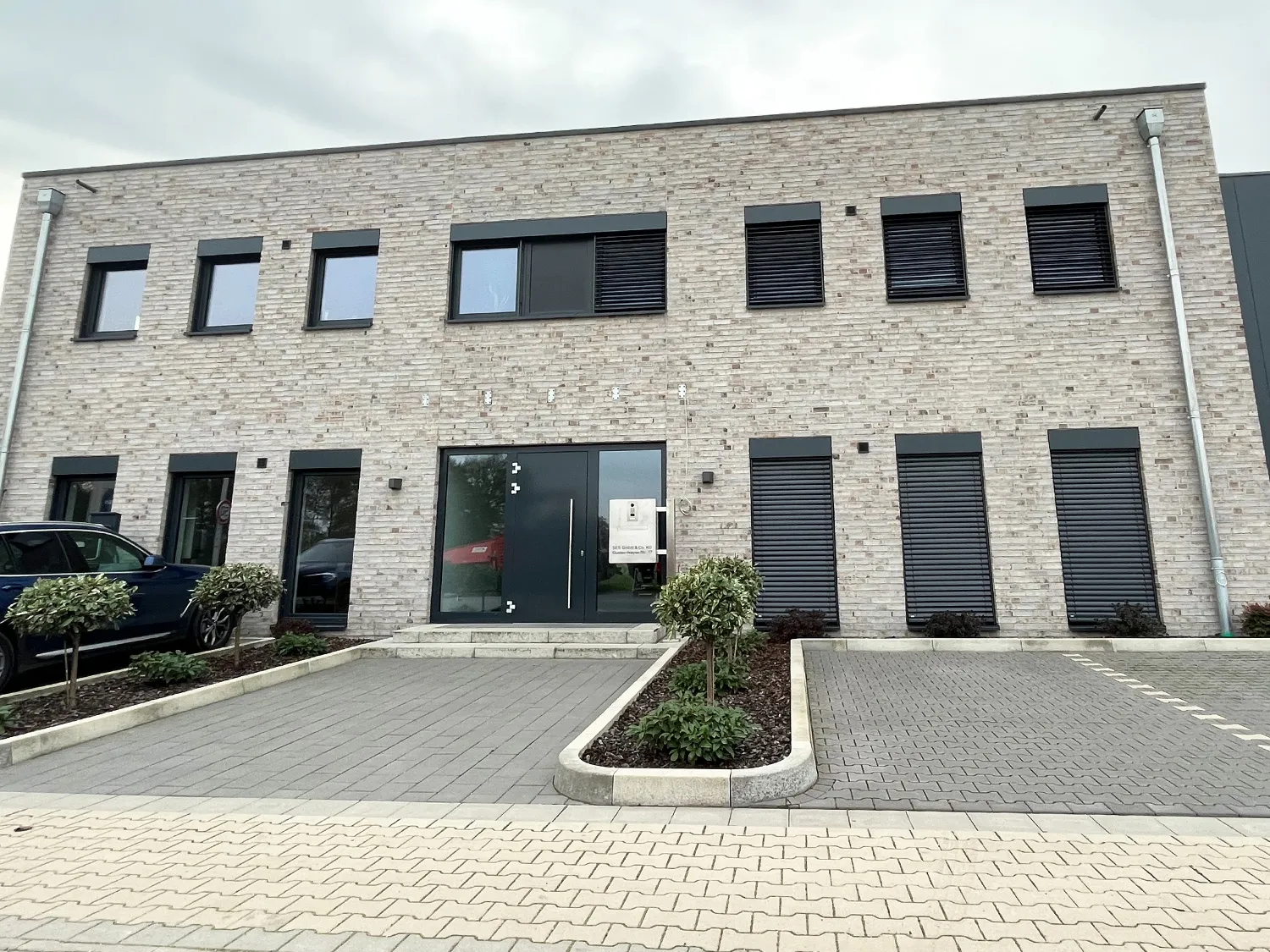 Modernes Gebäude mit heller Fassade, bodentiefen Glasfenstern. Davor ein gepflasterter Vorplatz