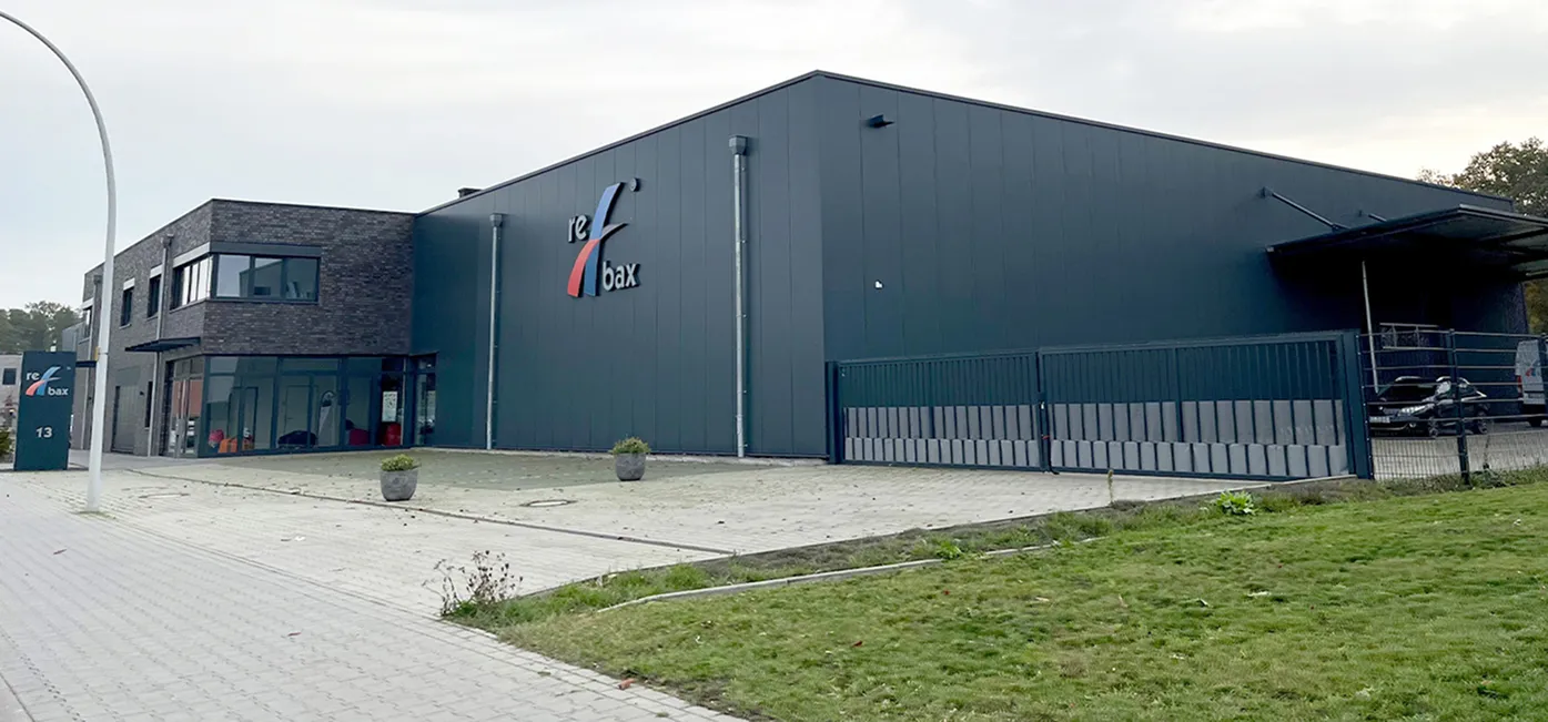 Außenansicht eines modernen Gewerbegebäudes mit dunkler Metall- und Klinkerfassade. An der Front befindet sich das Unternehmenslogo rebax, davor ein gepflasterter Vorplatz mit Pflanzkübeln und Einfahrtstor