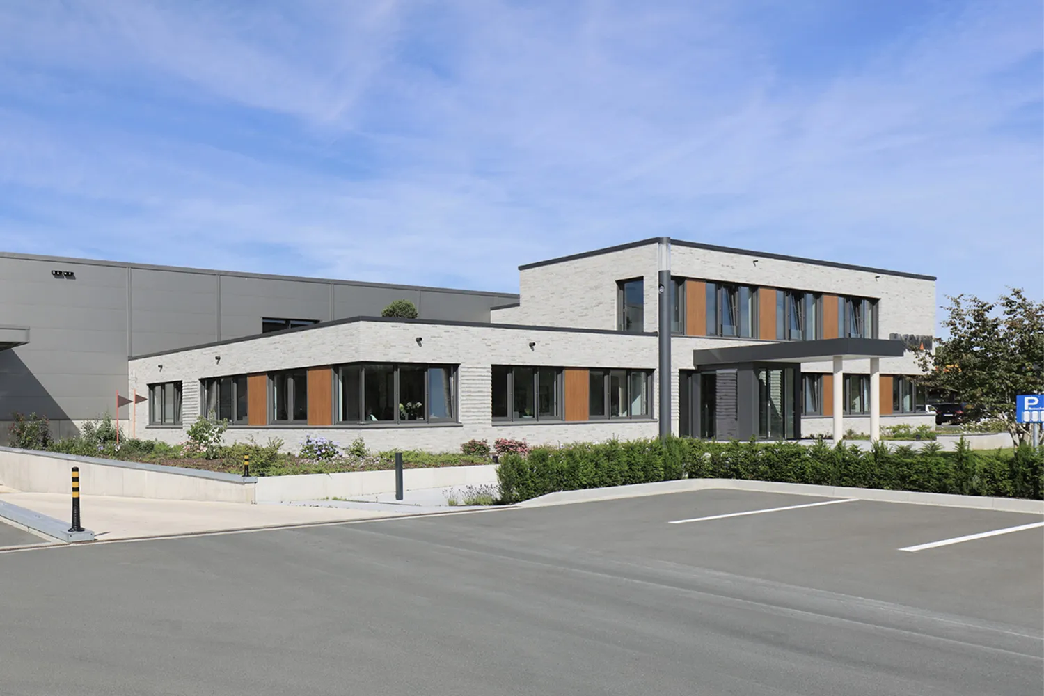 Modernes Firmengebäude mit heller Klinkerfassade, großen Fenstern und begrüntem Eingangsbereich.