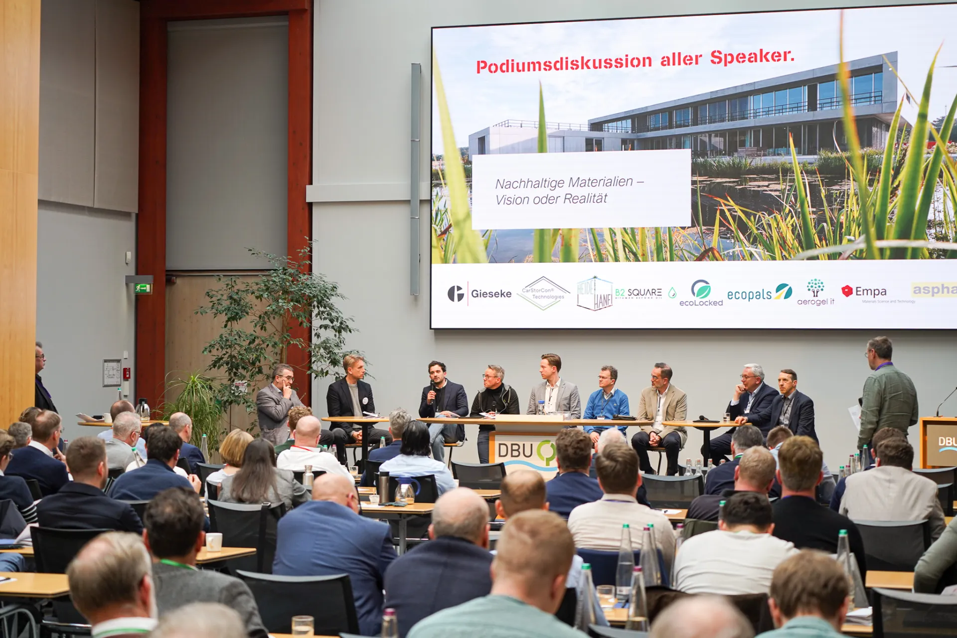 Podiumsdiskussion auf einer Fachveranstaltung zum Thema nachhaltige Materialien vor großem Publikum in einem Konferenzsaal.