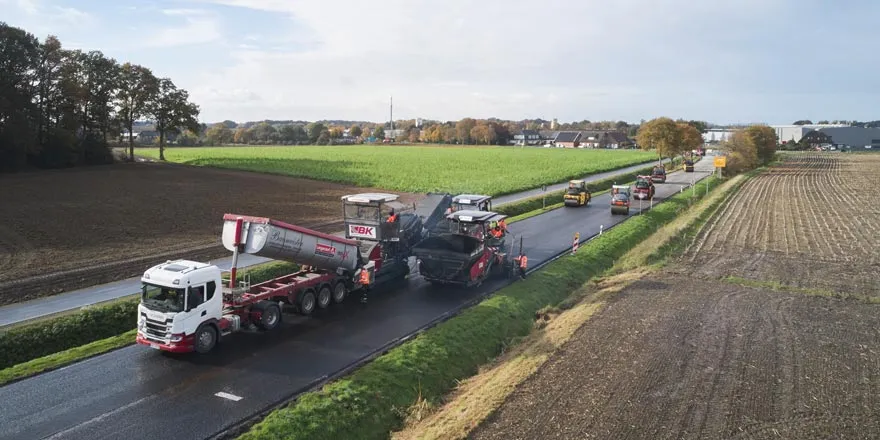 Straßenbau auf Landstraße mit Asphaltfertiger, Lkw und mehreren Baufahrzeugen zwischen Feldern aus der Vogelperspektive.