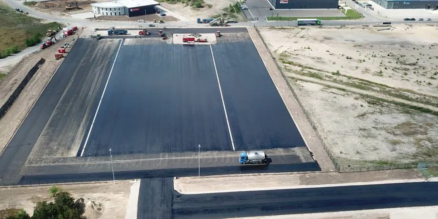 Großflächig asphaltierte Industrie- oder Verkehrsfläche aus der Luft, mit einzelnen Fahrzeugen und umgebendem Brachgelände.