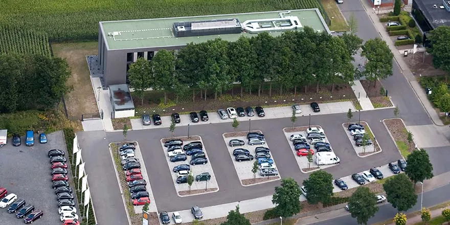 Luftaufnahme eines Verwaltungsgebäudes mit vorgelagerter Parkplatzanlage, Baumreihe und angrenzenden Feldern.