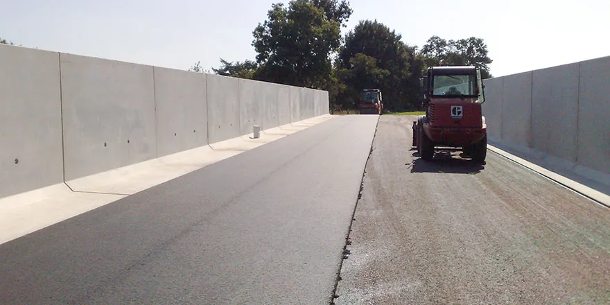 Frisch asphaltierte Fahrbahn zwischen hohen Betonwänden, rechts daneben eine kleine Walze auf der Baustelle.