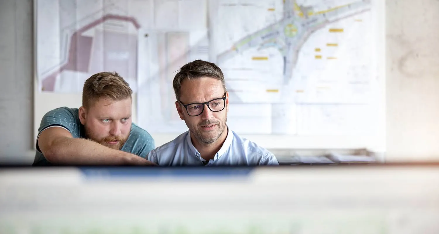 Zwei Mitarbeiter in der Verwaltung vor einem Bildschirm und Bauplan im Hintergrund