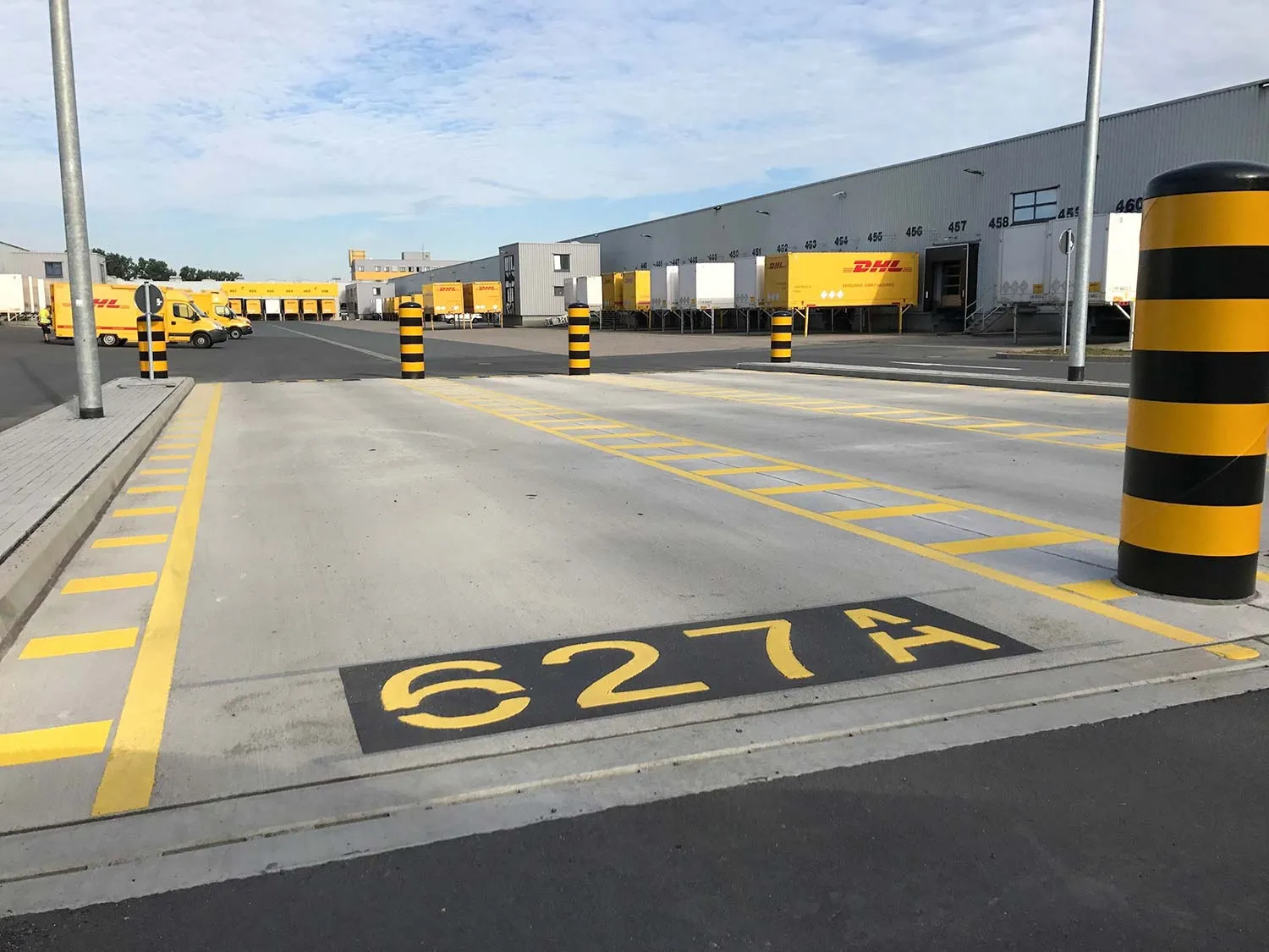 Verkehrsfläche an einer DHL-Verladehalle mit Fahrbahnmarkierungen, Pollern und angedockten Fahrzeugen.
