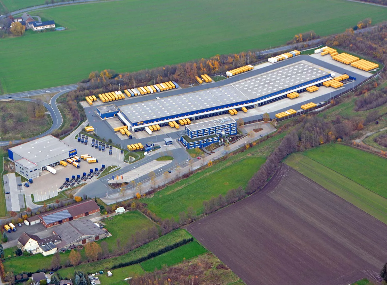 Luftaufnahme eines Logistikstandorts mit Hallengebäude, Verwaltungsbau und abgestellten gelben Wechselbrücken.