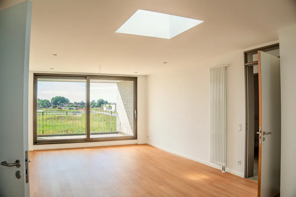 Heller, leerer Wohnraum mit Holzfußboden, großem Fenster und Blick ins Grüne.