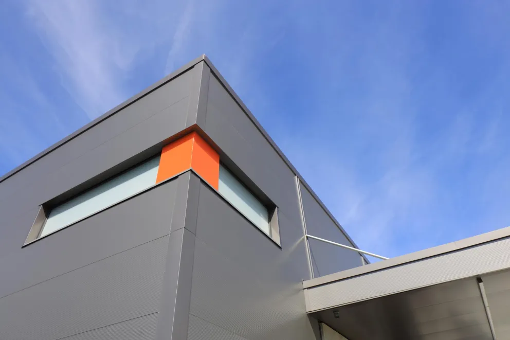 Eingangsbereich eines modernen Bürogebäudes mit heller Klinkerfassade, großen Fensterflächen und bepflanztem Vorbereich.