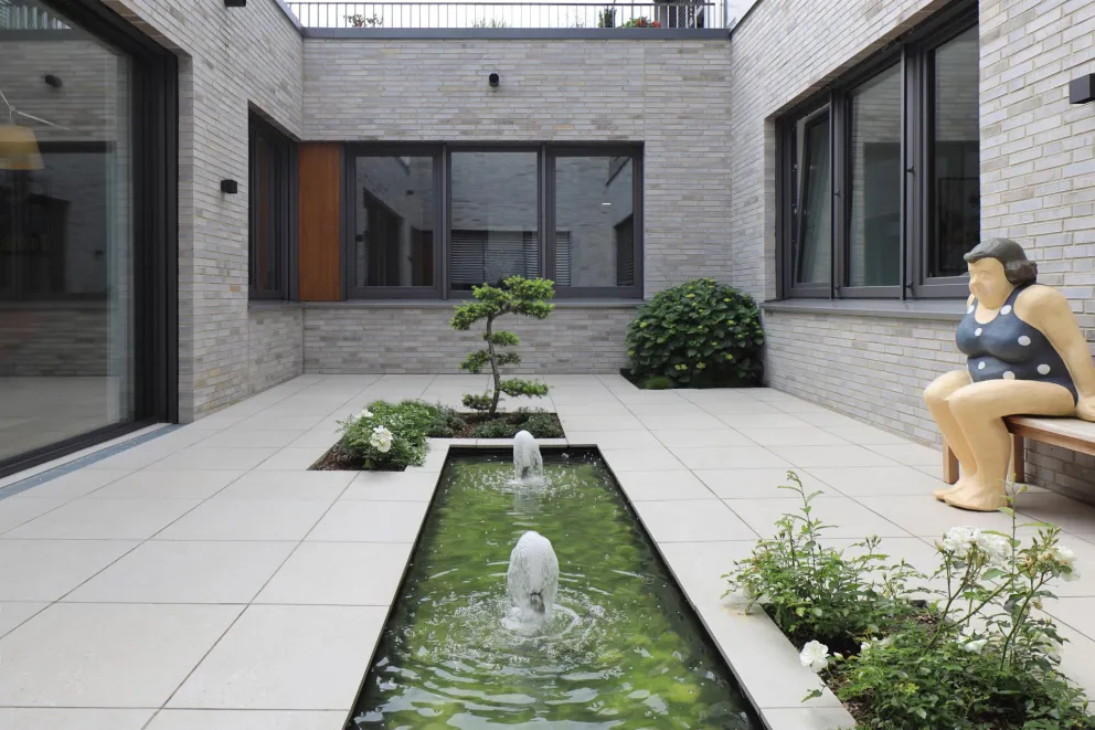 Ruhiger Innenhof mit langem Wasserbecken, bepflanzten Flächen und Sitzfigur in moderner Architektur.